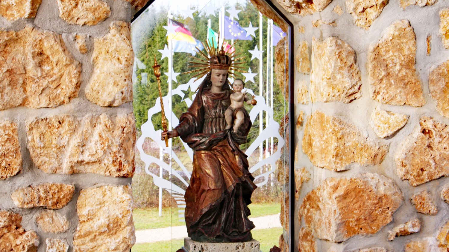 260915gnadenweiler (06a).JPG - Die barocke Muttergottes (Gnadenbild): Das Herzstück der Kapelle ist eine historische, ca. 125 cm hohe Muttergottesfigur aus der Schweiz, die um das Jahr 1750 im Barockstil gefertigt wurde. Sie verbindet die moderne Architektur des Raumes mit der Jahrhunderte alten Tradition der Marienverehrung. Im Fenster eingraviert : 12 Sterne – verweisen auf Offenbarung 12,1"Und es erschien ein großes Zeichen im Himmel: eine Frau, mit der Sonne bekleidet, und der Mond unter ihren Füßen und auf ihrem Haupt eine Krone von zwölf Sternen"In der katholischen Tradition wird diese Bibelstelle auf Maria hin interpretiert.Hier kann man auch eine Querverbindung zur Europafahne sehen - auf diesem Foto flattert sie draußen am Fahnenmast - die ebenfalls einen Kreis von 12 Sternen hat. Hier wird die Zwölf als Symbol der Vollkommenheit und Vollständigkeit gedeutet.Auch im Fenster eingraviert: eine Jakobsmuschel – weist auf den Beuroner Jakobsweg hin, der hier an der Kapelle vorbei von Hechingen nach Messkirch führt.